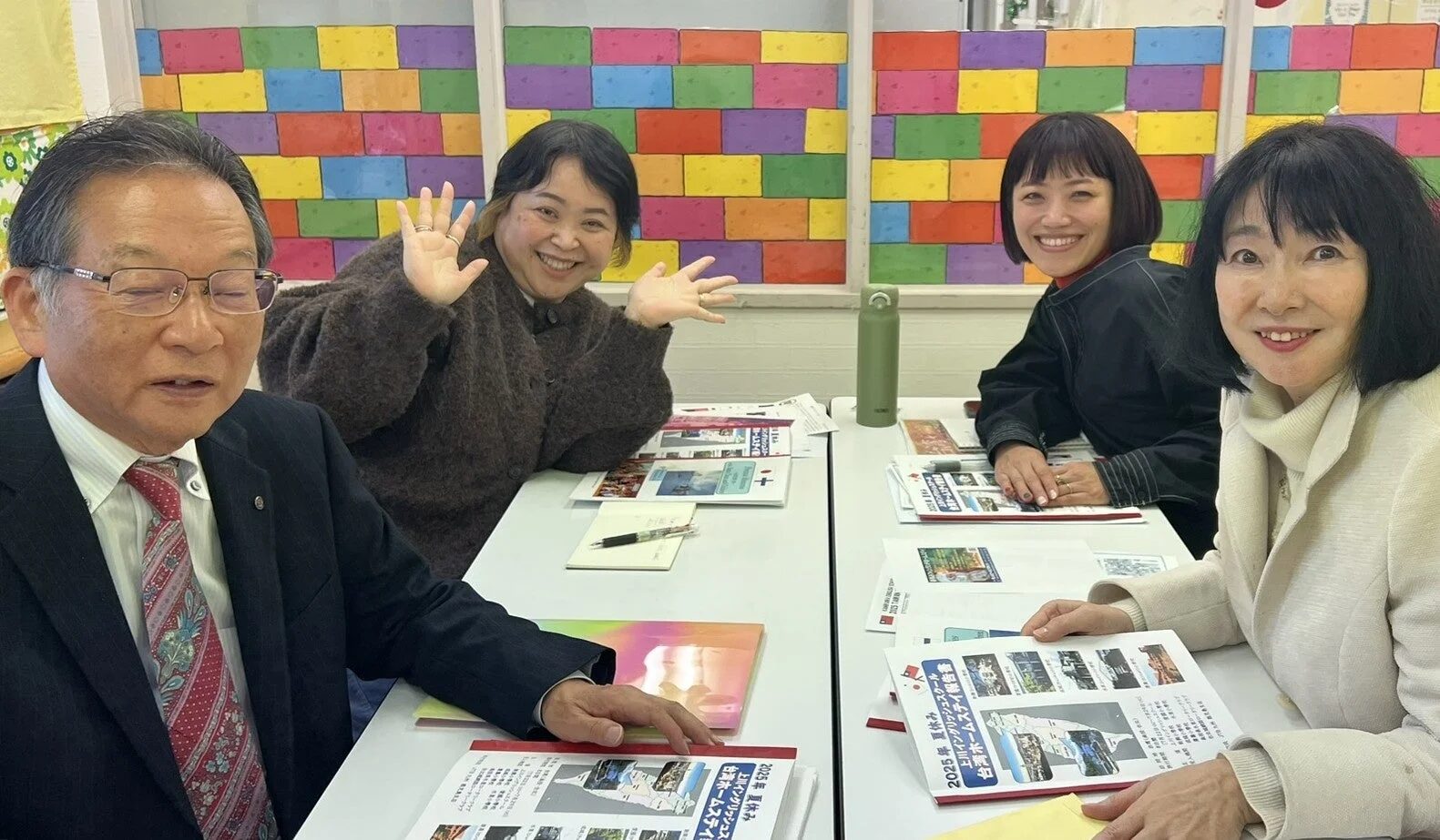 カラフルな壁を背景に、4人の男女がテーブルを囲み、笑顔で会話している。手元には「2025年夏休み 台湾ホームステイ報告書」などの書類があり、和やかな雰囲気で打ち合わせや報告会が行われている様子がうかがえる。