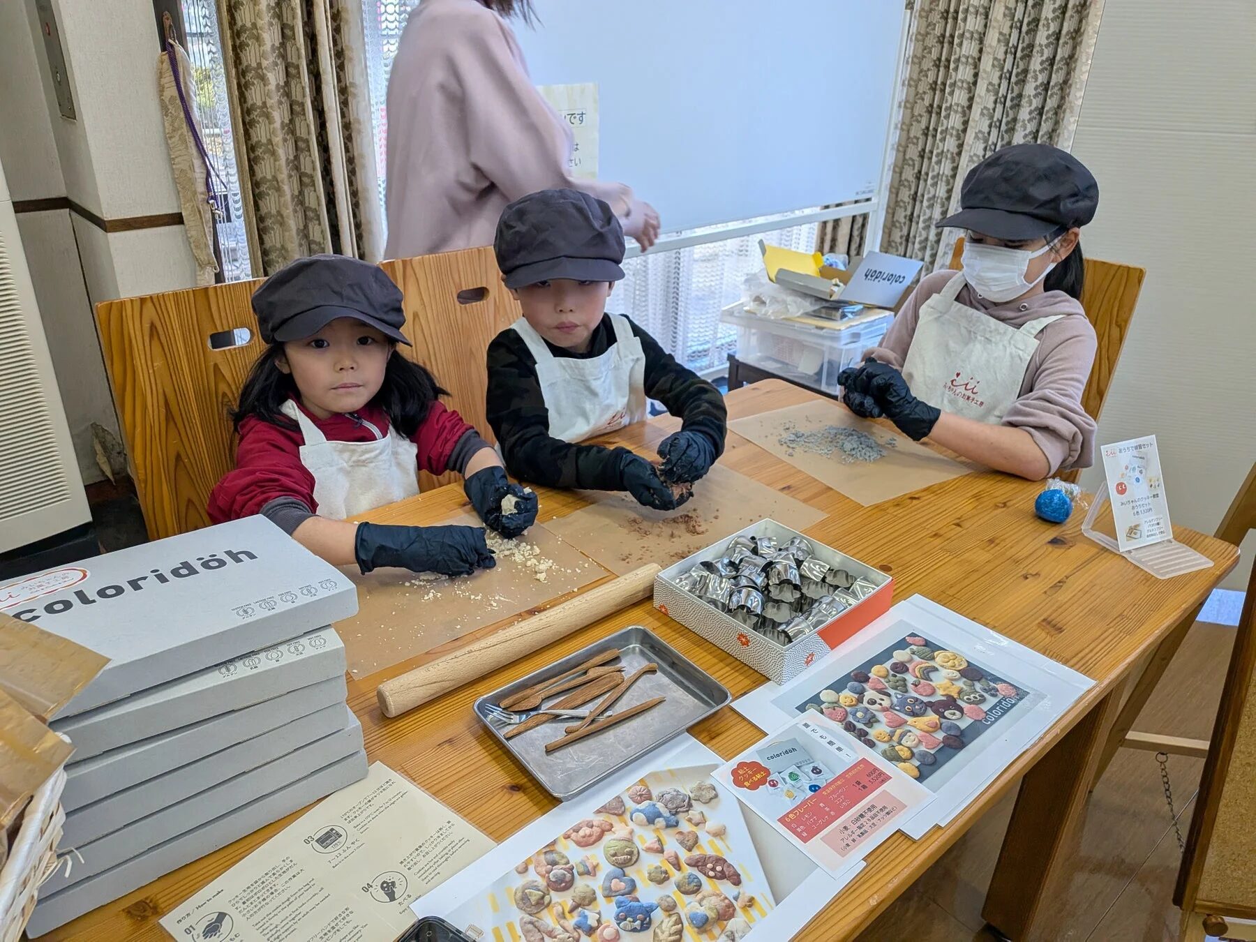 子供たちがエプロンと手袋を着用し、テーブルで粘土のような素材を使ってクッキー作りまたは造形活動をしている様子